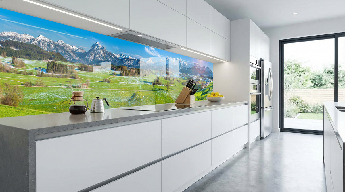 Moderne weiße Küche mit der Küchenrückwand „Sonnenaufgang Alpenwiesen“ von glasposter.com und großen Fenstern.